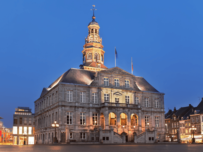 The Maastricht City Hall illuminated in the evening, featuring a grand clock tower and a symmetrical facade.