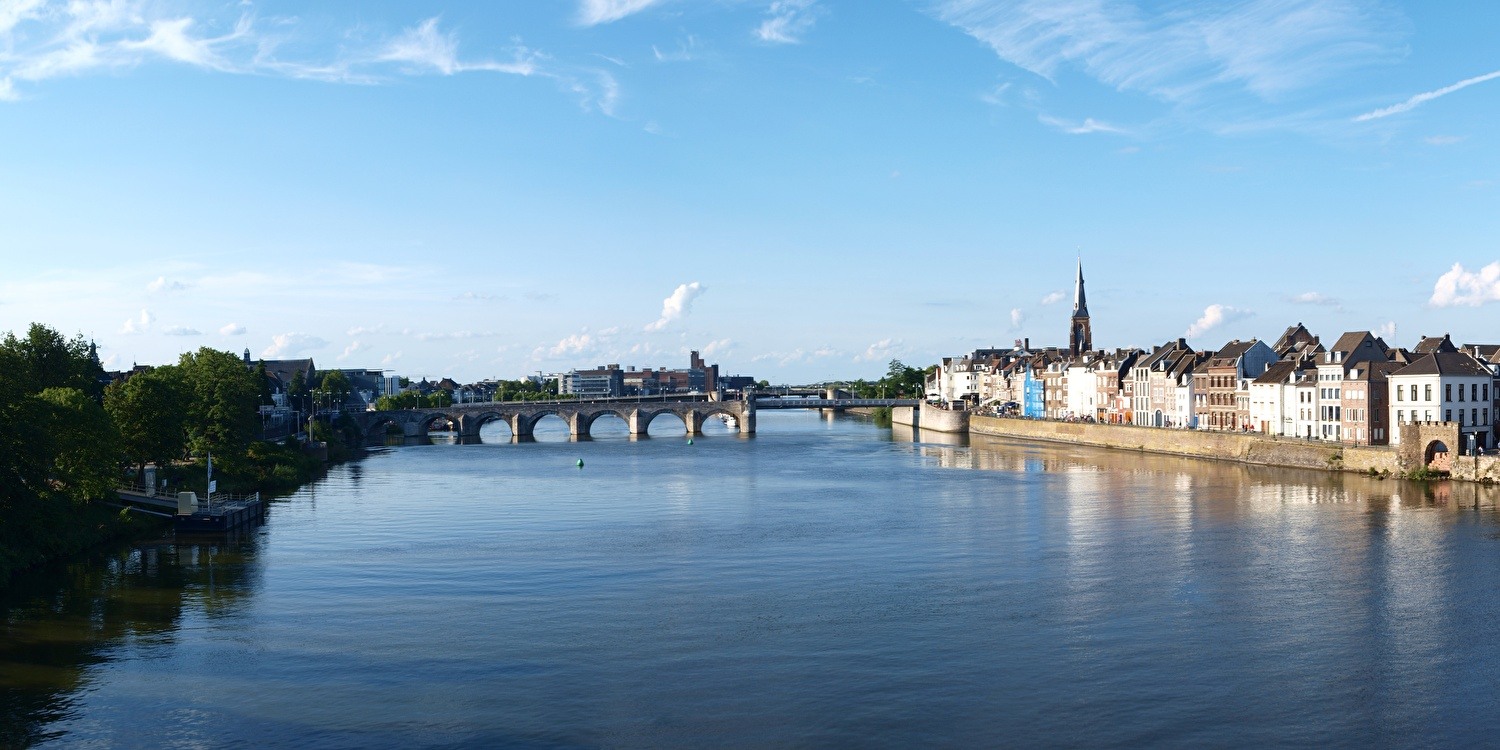 Panorama van Maastricht met uitzicht op de Maas, de iconische Sint-Servaasbrug en de historische gevels langs de kade onder een blauwe lucht.